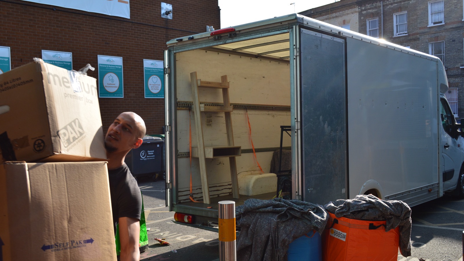 Mover carrying packed cardboard boxes beside an open Luton removal van during an operational job