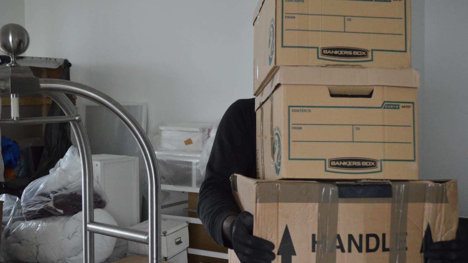 Removal worker carrying three sealed cardboard boxes beside a trolley inside a property