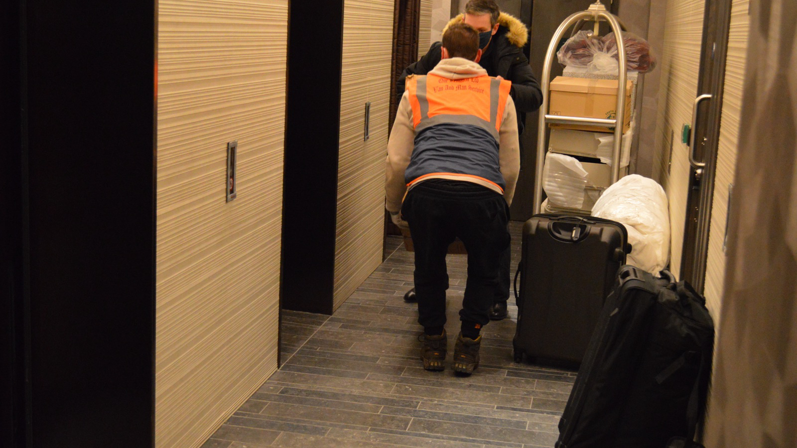 Two movers using a trolley with boxes and luggage in a managed apartment corridor