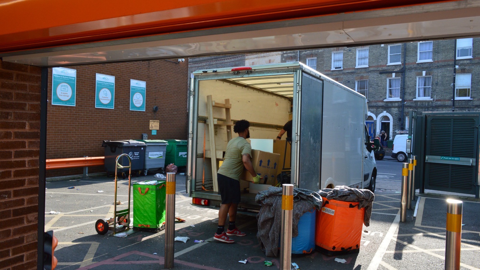 Removal worker loading boxed goods beside a trolley, blankets and an open Luton van