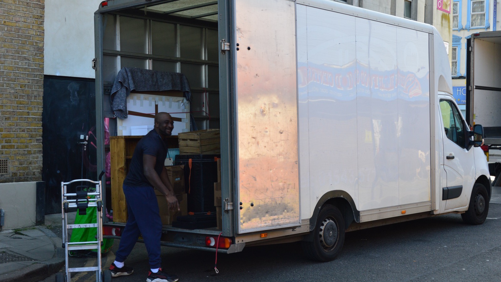 Unbranded removal mover with a hand trolley beside a white Luton van on a tight London street